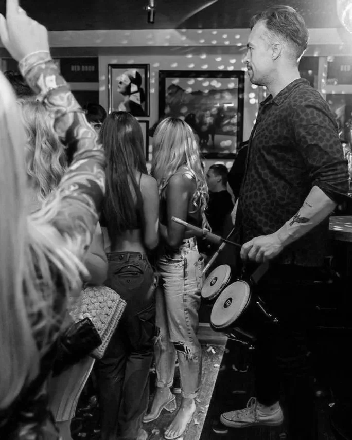 A black and white photo of a crowded indoor party, with a man holding a percussion instrument, possibly a drum, among young women with long hair. The background shows party lights and framed pictures on the wall.