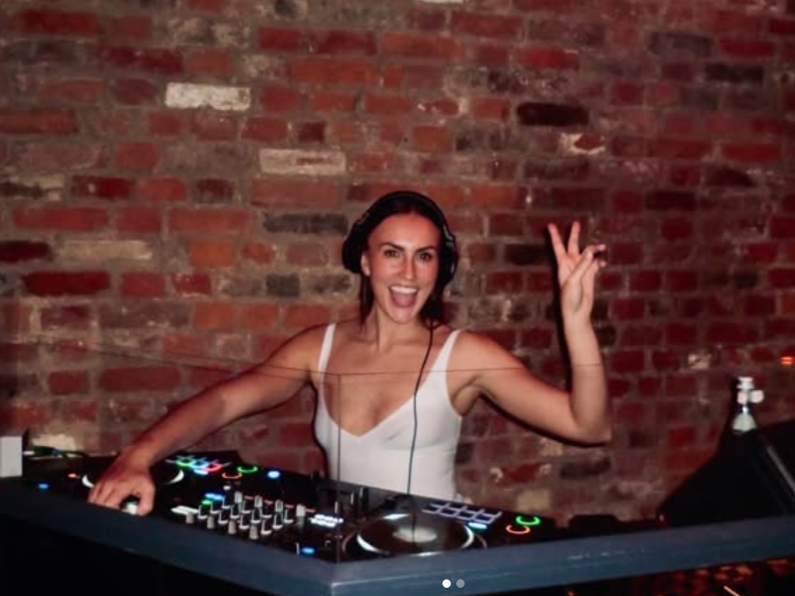A woman DJ wearing headphones and a white tank top, smiling and making a peace sign with her right hand behind a DJ mixing console in front of a brick wall.