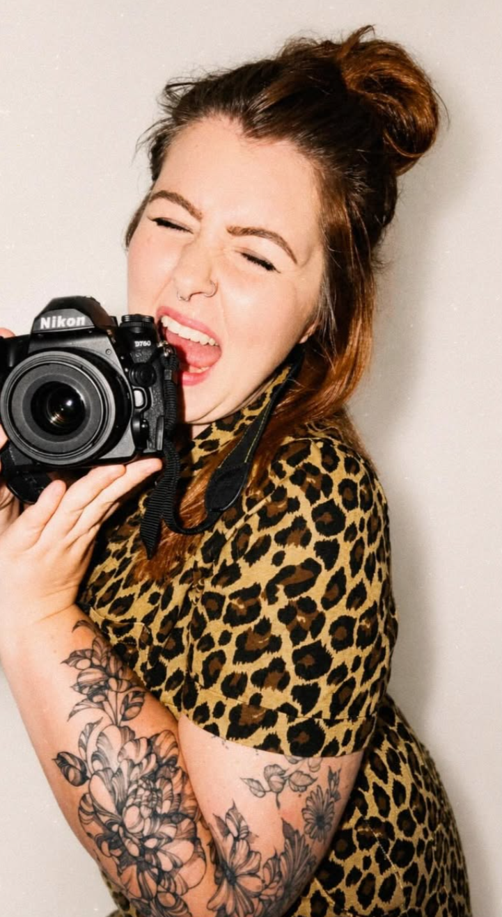 A woman with reddish-brown hair in a bun, wearing a leopard print shirt, holding a Nikon camera, with tattoos on her arm, making a playful expression with her tongue out and eyes closed.