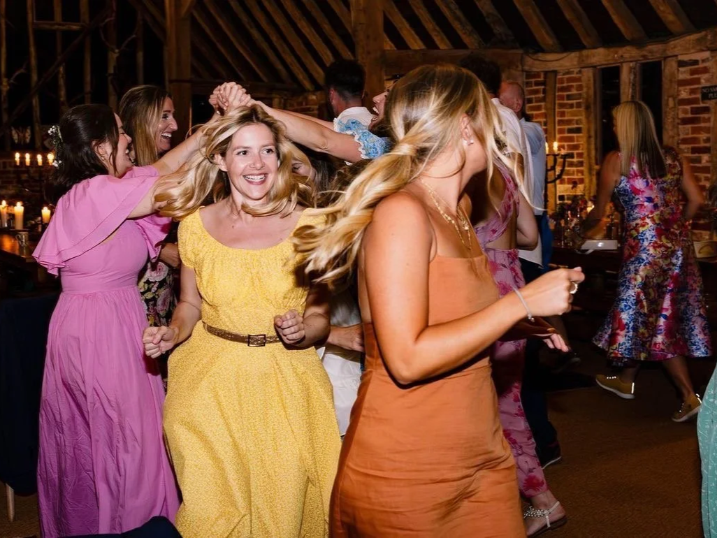 Group of women dancing and smiling at a lively party in a rustic venue with exposed brick walls and wooden beams, candles and decorations in the background.