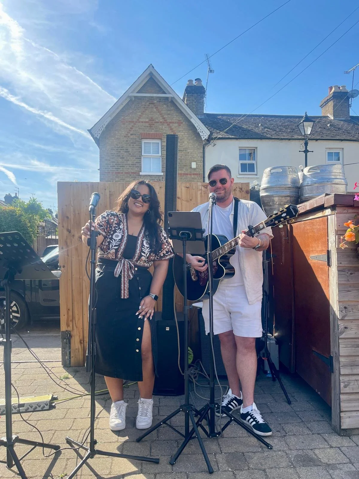 Two musicians performing outdoors, one woman singing into a microphone and holding a microphone handle, and one man playing an acoustic guitar and singing into a microphone, both wearing sunglasses, with a wooden fence and a brick house in the background.