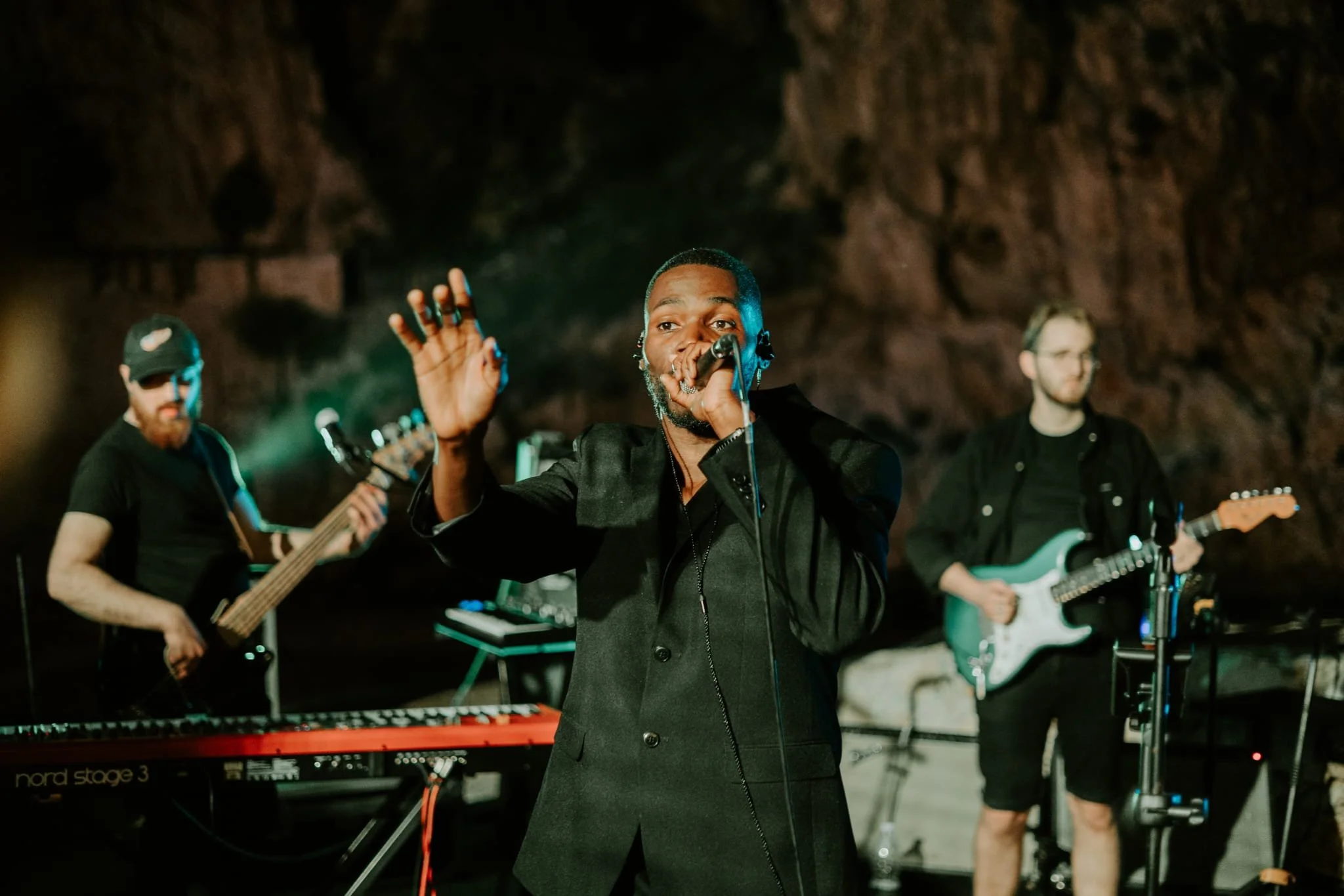 A band performs in a dark, rocky setting. A man in the center sings into a microphone with his hand raised. Two guitarists and a keyboardist accompany him in the background.