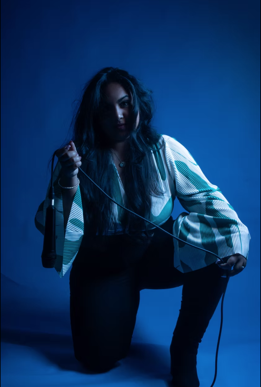 A woman with dark hair kneeling on the floor holding a microphone with a blue background.