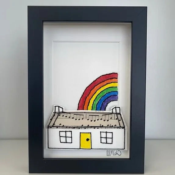 Handmade card frame containing a drawing of a house with a yellow door and a rainbow in the background.