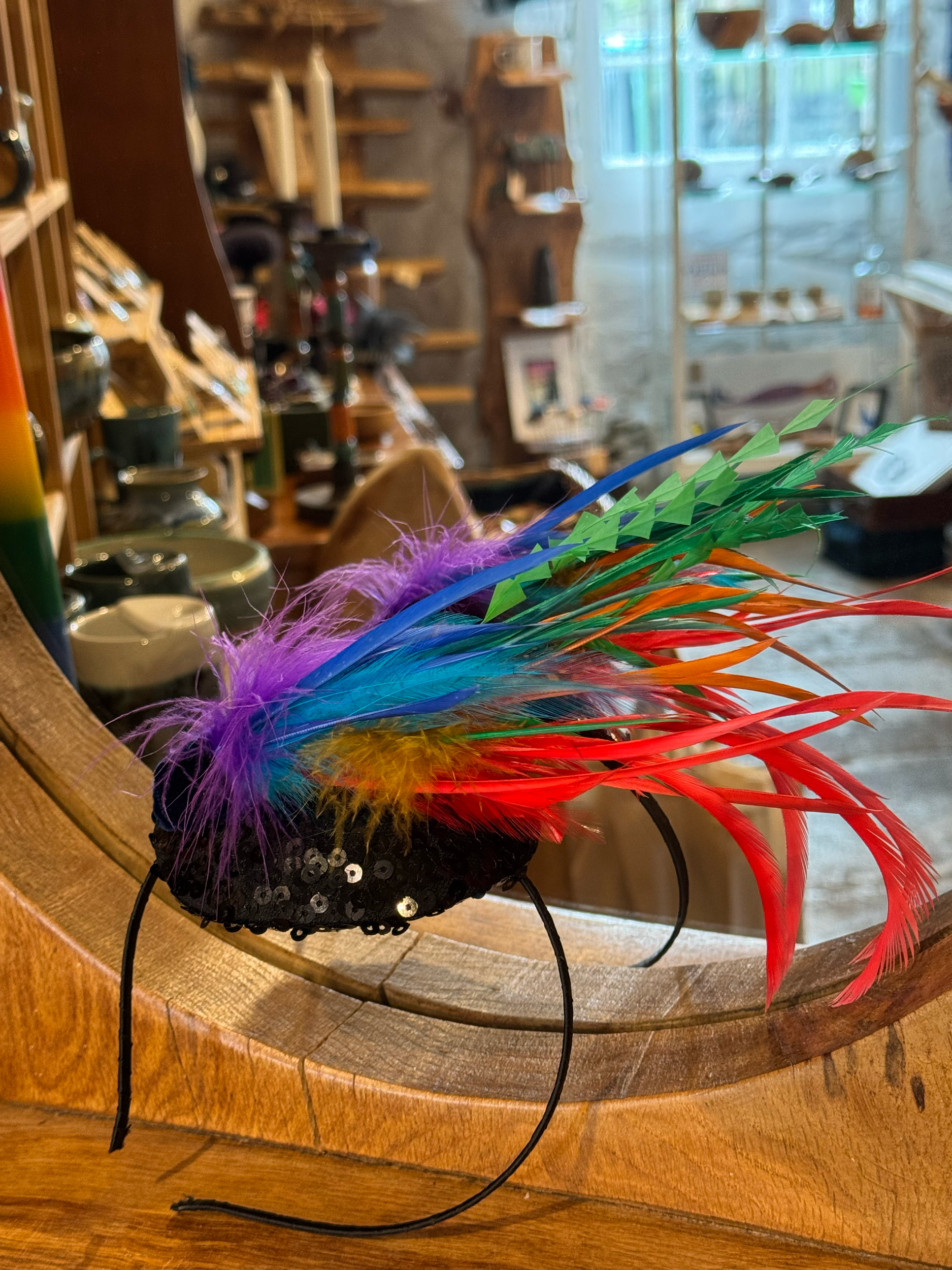 A colorful headband decorated with vibrant feathers in red, purple, blue, green, and orange, and sequins, placed on a wooden table inside a craft or gift shop.