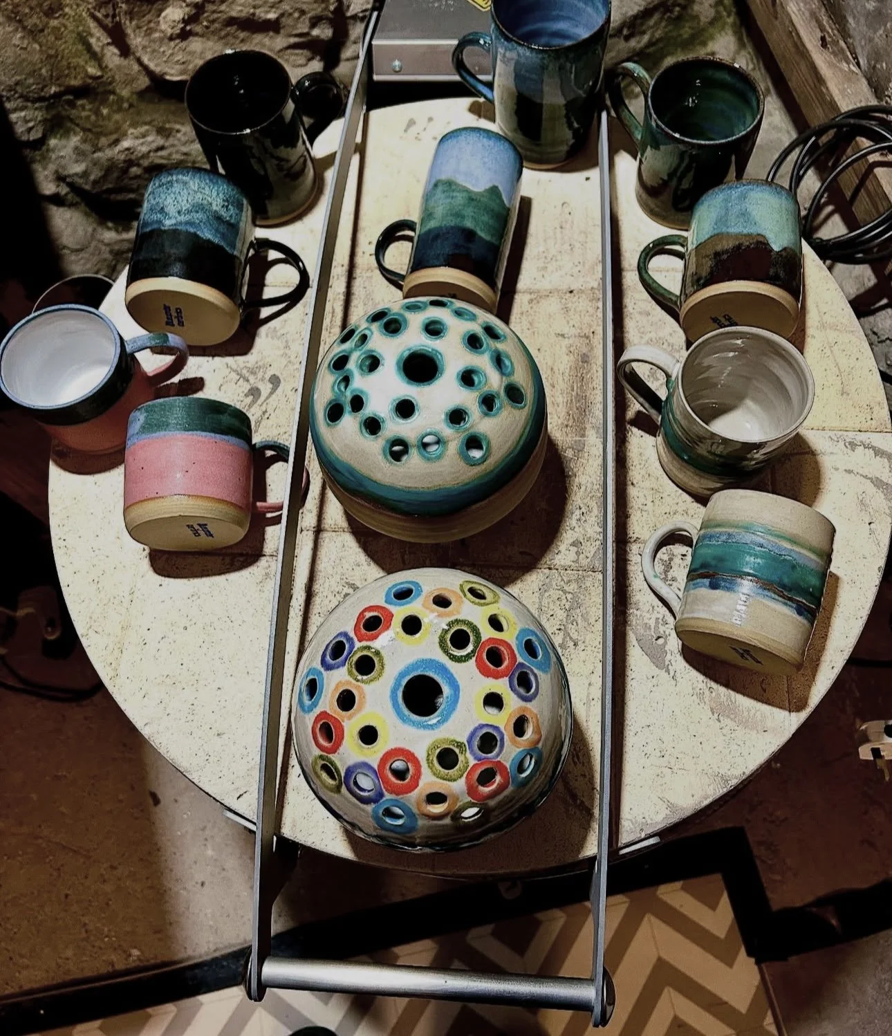 Colorful ceramic mugs and bowls with dotted and striped designs arranged on a rustic round wooden table.
