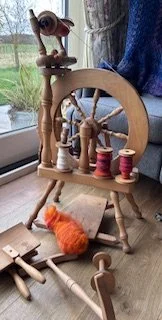 A wooden dollhouse with miniature furniture and accessories, including a sewing machine, a spool of red thread, a small orange fluffy toy, and other miniature items, placed on a table near a window.