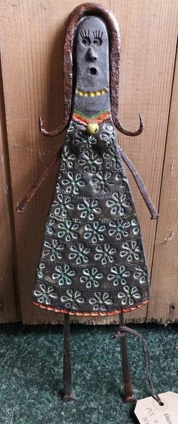Rustic metal sculpture of a woman wearing a floral-patterned dress, necklace, and with curly hair, standing against a wooden background.