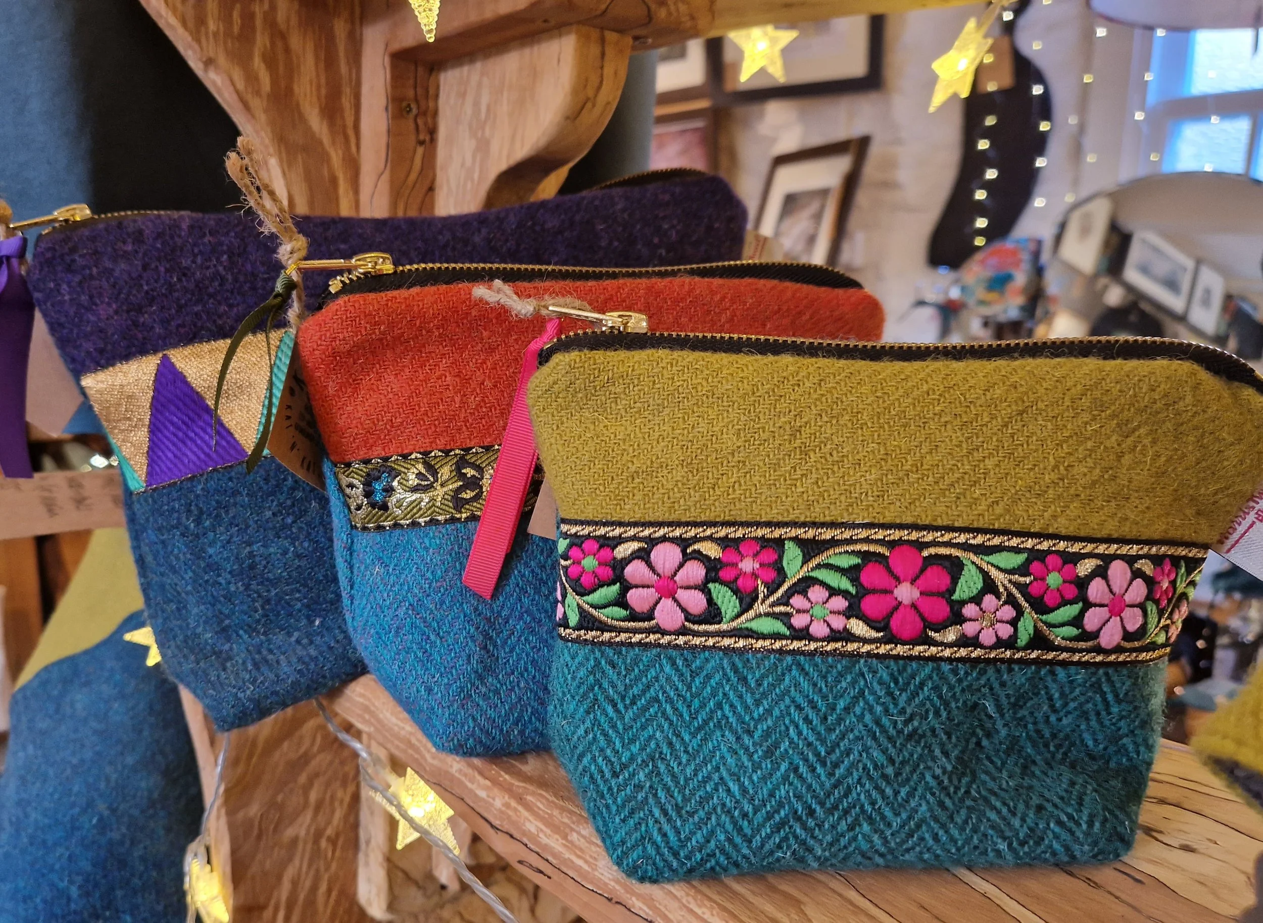 Colorful woven pouches with embroidered floral designs, displayed on a wooden shelf in a store with string lights and framed pictures in the background.