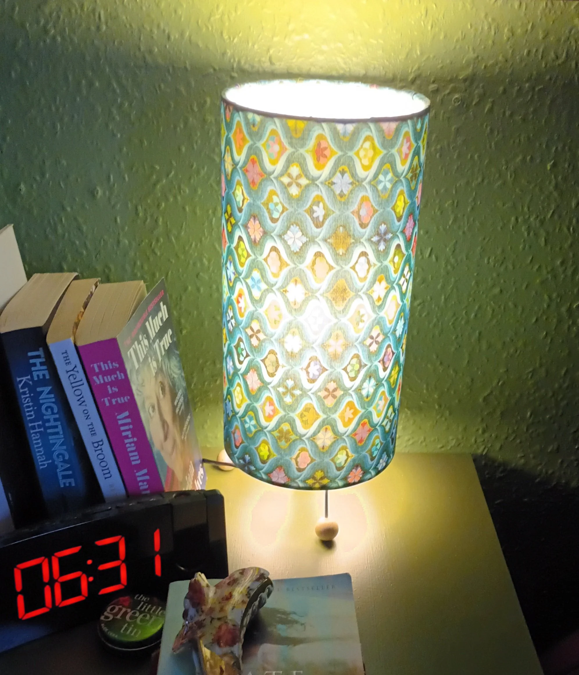 A colorful bedside lamp with a patterned shade on a nightstand, next to a stack of books and an alarm clock displaying 06:31. There is a small decorative item and a tin can on the table as well.