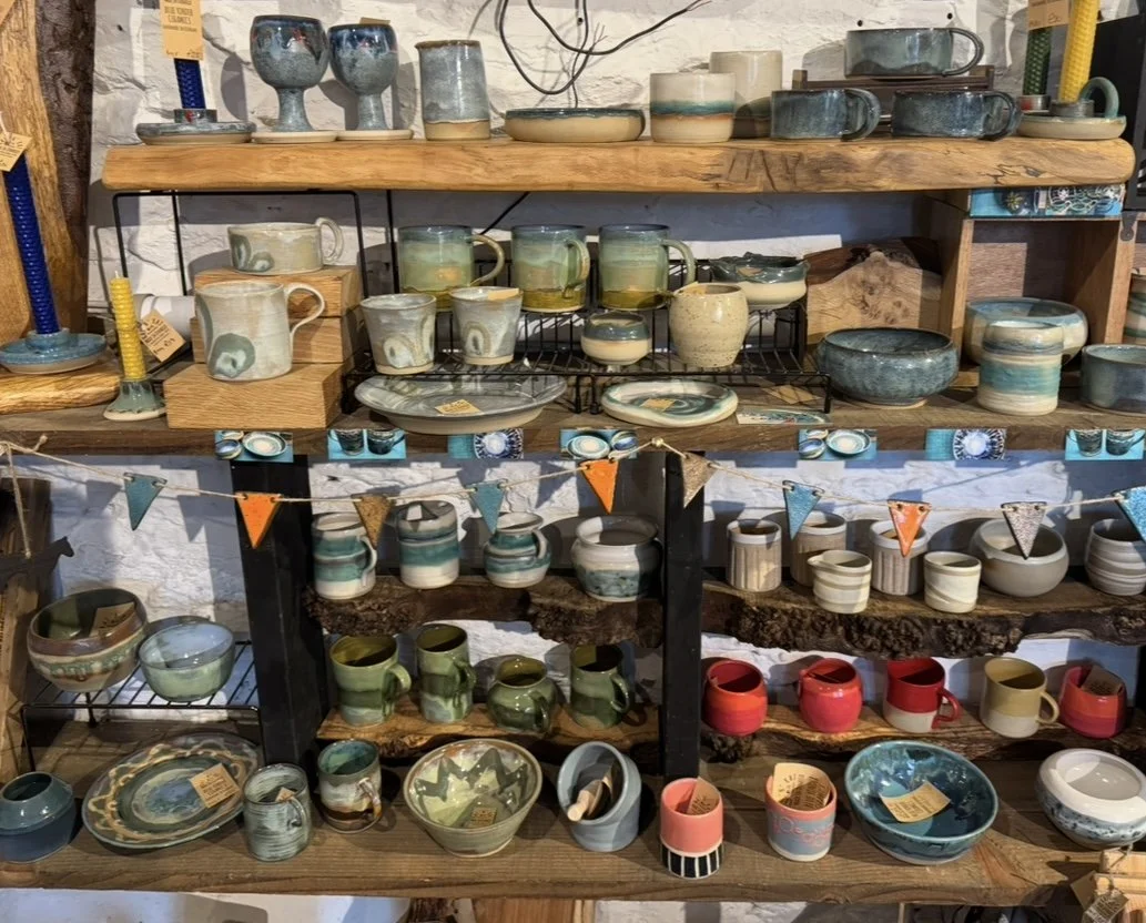Display of various handmade ceramic pottery, including mugs, bowls, and plates, arranged on wooden and metal shelves in a shop. Some items are on the top shelf, and others are grouped on lower shelves, with small flags and tags attached.