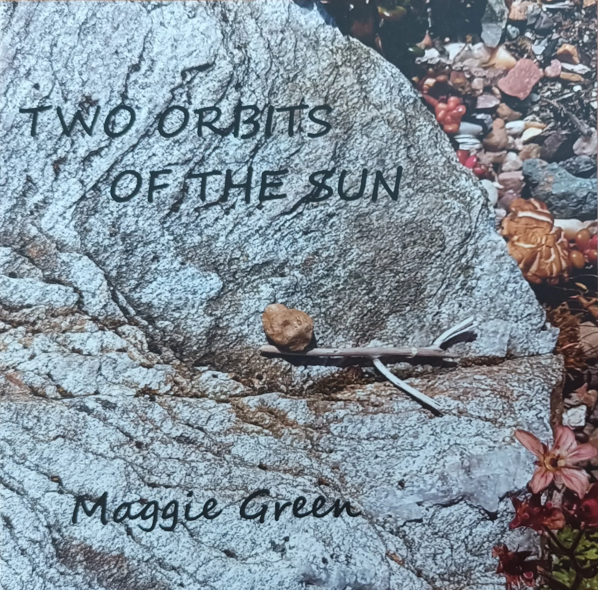 A large light gray granite rock with the quote 'Two orbits of the sun' and the author's name, Maggie Green, etched into it. The rock is surrounded by small pebbles and some reddish-brown flowers at the bottom right corner.