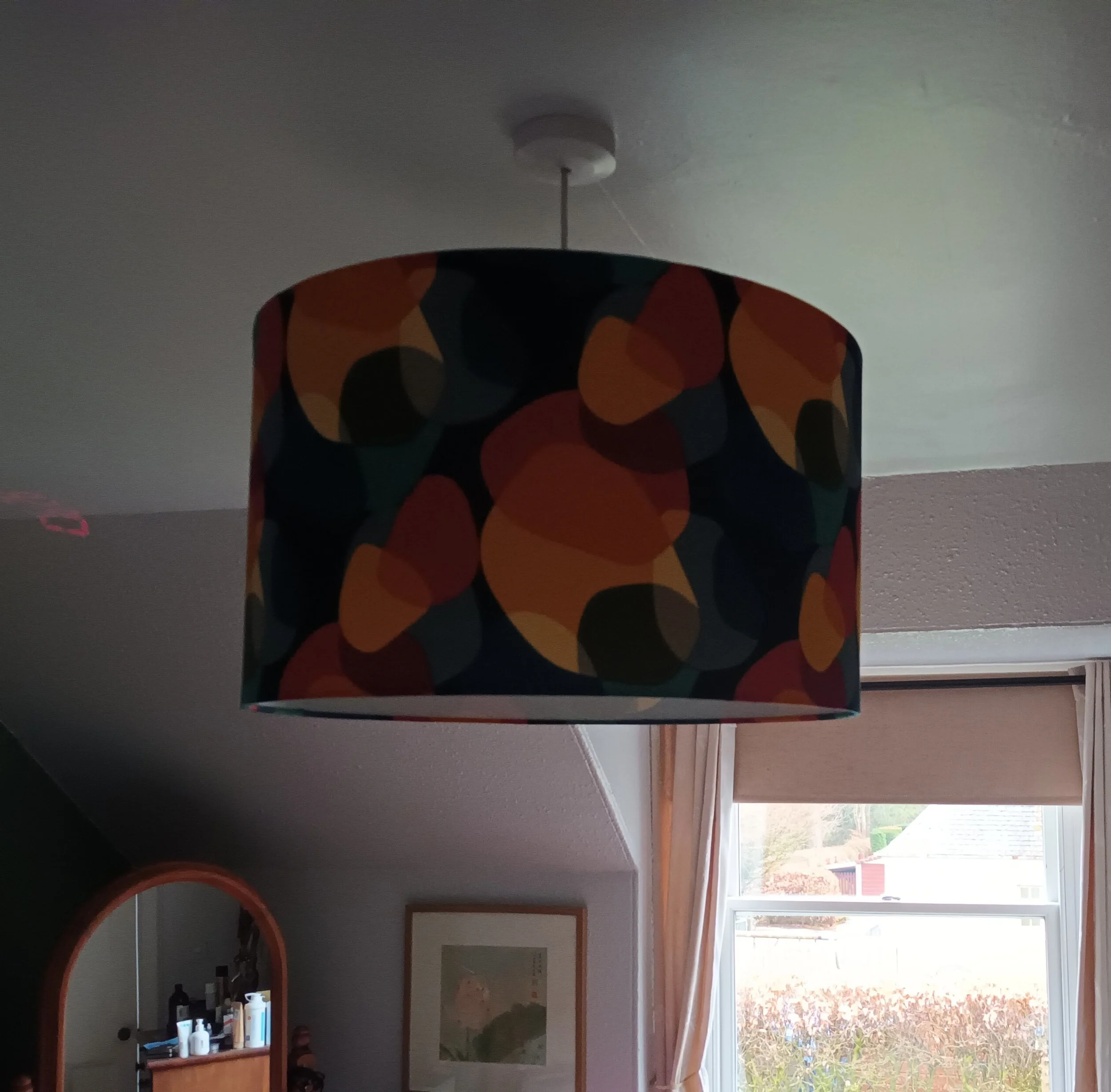 Colorful patterned lampshade hanging from the ceiling in a room with a window, mirror, and framed picture on the wall.