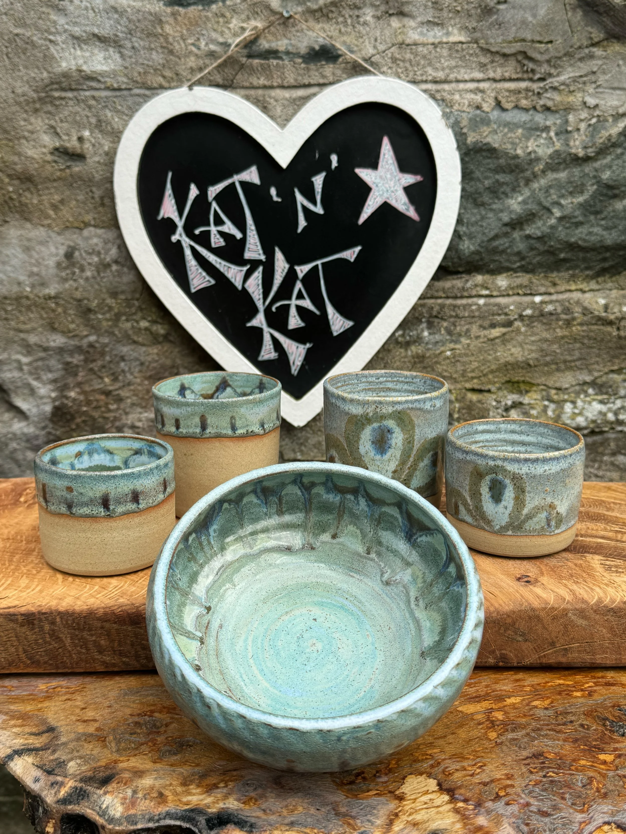 Set of five handcrafted ceramic bowls with blue and beige glaze, displayed on a wooden surface against a stone wall background with a heart-shaped chalkboard that reads 'KAT N' on top.