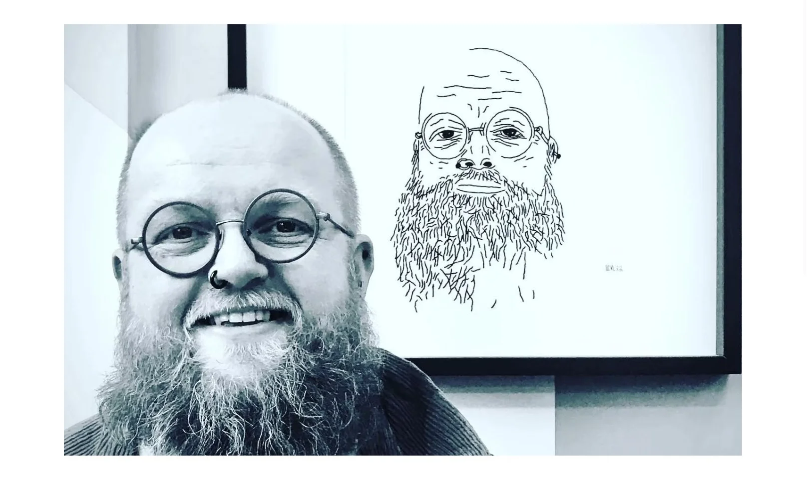 A smiling man with a beard, glasses, and a nose ring standing indoors in front of a framed line drawing of a bearded man with glasses.