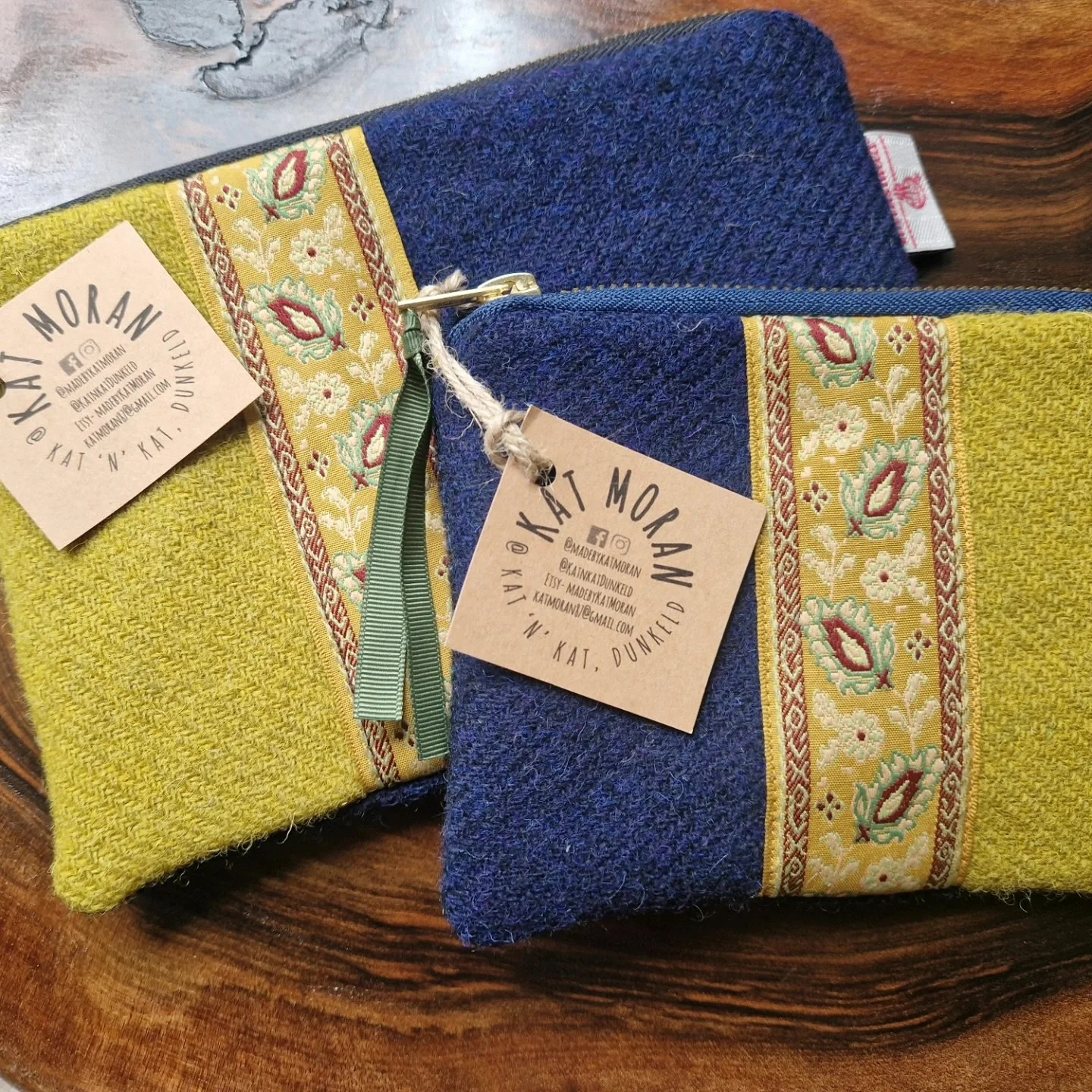 Two handmade fabric pouches with a floral embroidered trim, one in yellow and the other in dark blue, placed on a wooden surface. Each pouch has a beige tag with the name 'Kait N Kat' and social media handles.