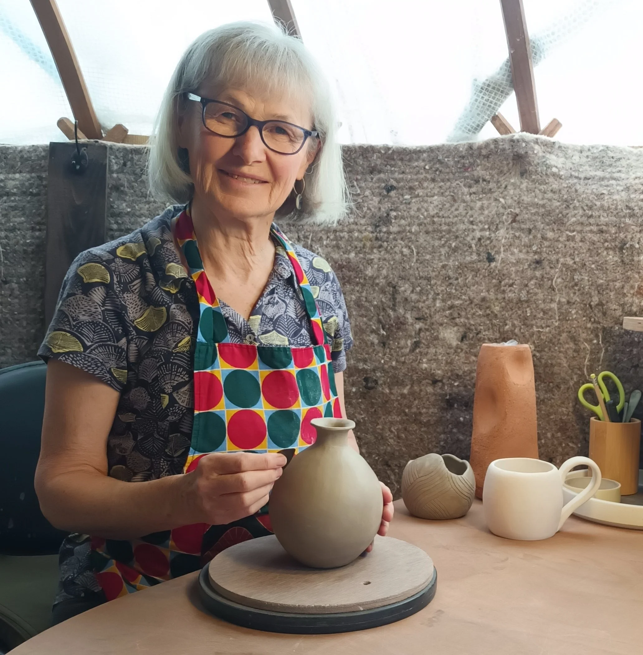 An elderly woman with short white hair, glasses, and a patterned shirt, wearing a colorful apron with circles, is shaping a clay vase in pottery studio. She is smiling and holding the vase with both hands, standing by a table with pottery tools and finished ceramic pieces.
