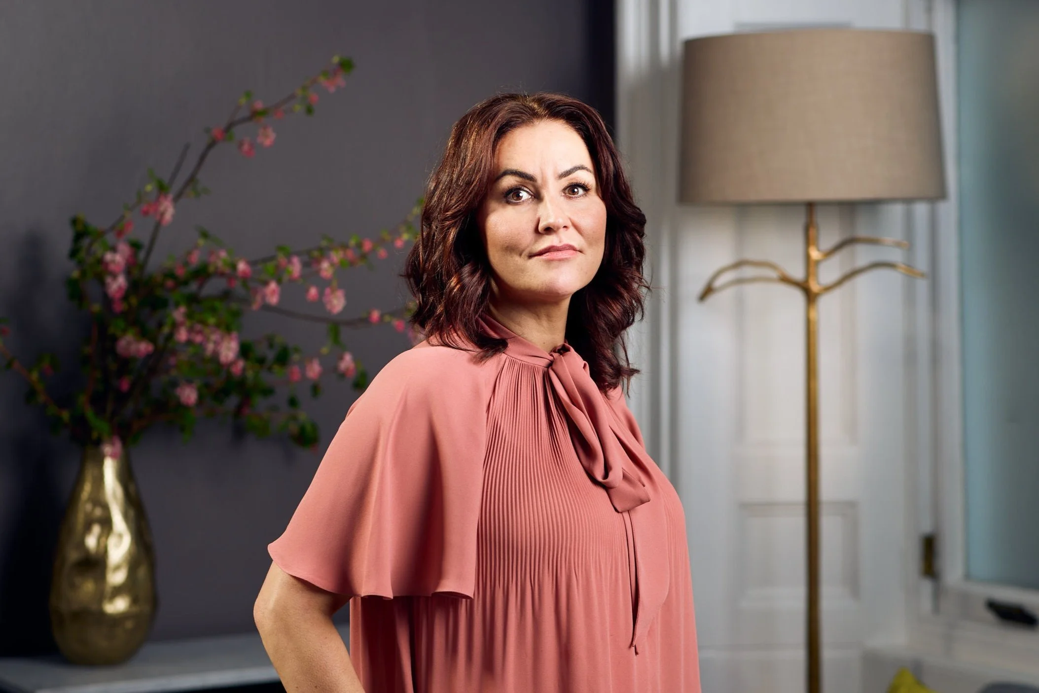 A woman with shoulder-length curly brown hair wearing a pink dress with a bow tie neckline standing indoors near a white door and a gold-coated floor lamp, with a dark wall and a vase of pink blossoms in the background.