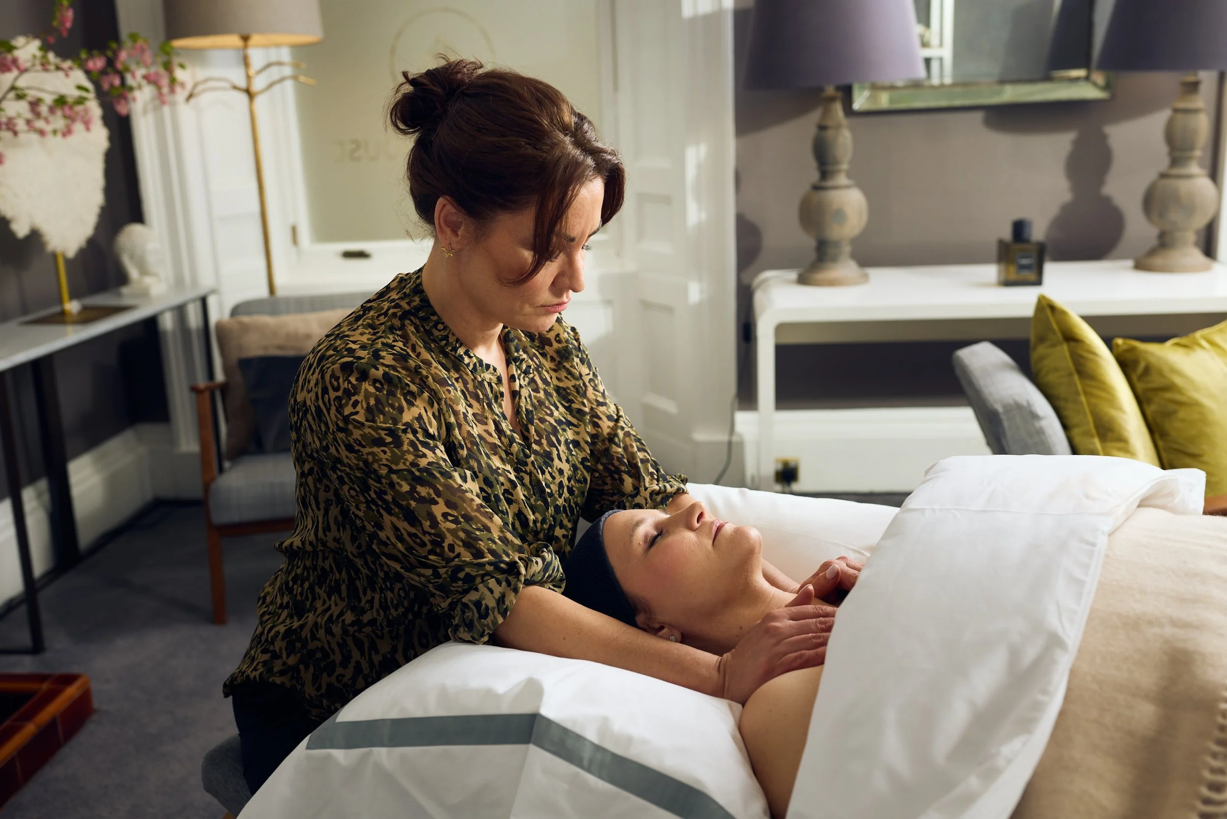 A woman giving a massage to another woman lying on a bed in a well-decorated room with lamps and pillows.