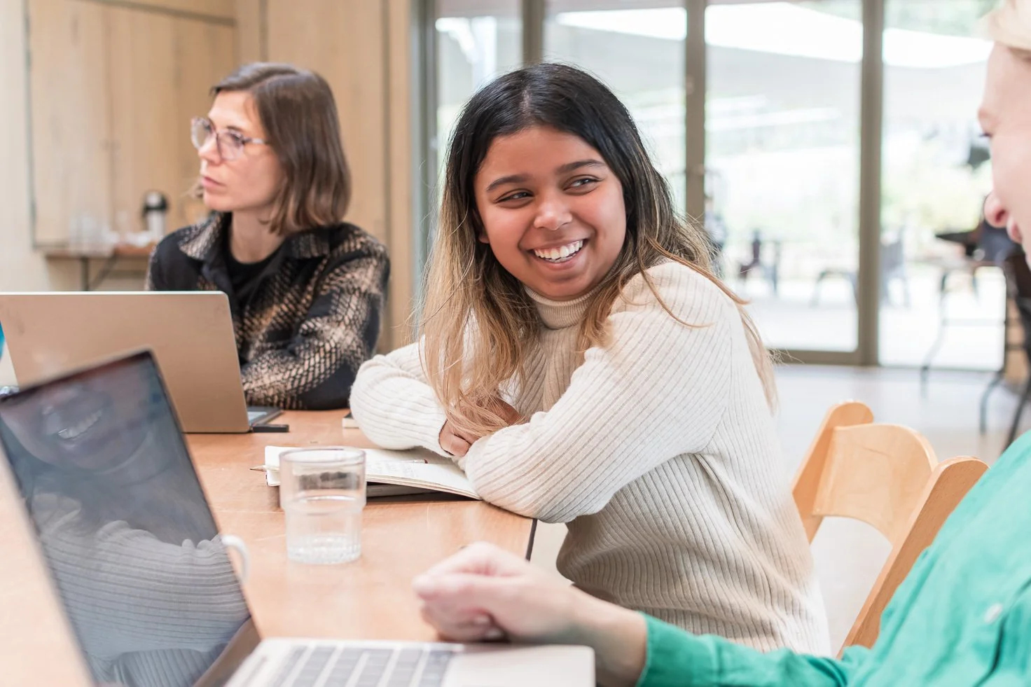 A group of women sitting at a table with laptops and notebooks, engaging in a discussion in a bright, modern space.