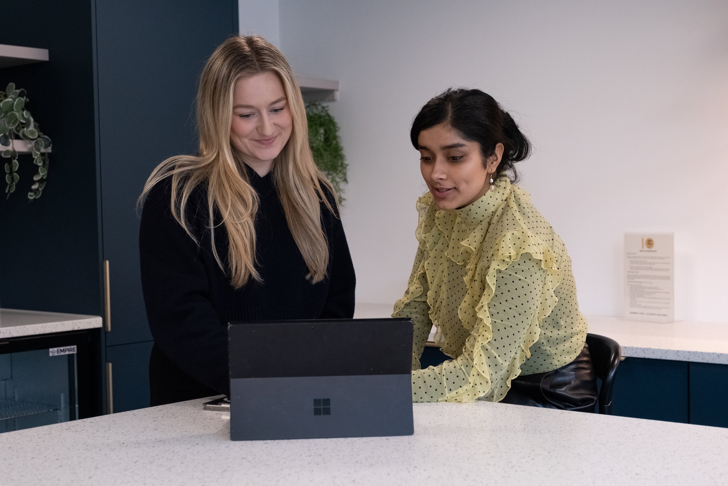 Two women are standing at a counter looking at a laptop screen. One has long blonde hair and is wearing a black top, while the other has dark hair styled in loose waves and is wearing a yellow polka-dot blouse with ruffles.