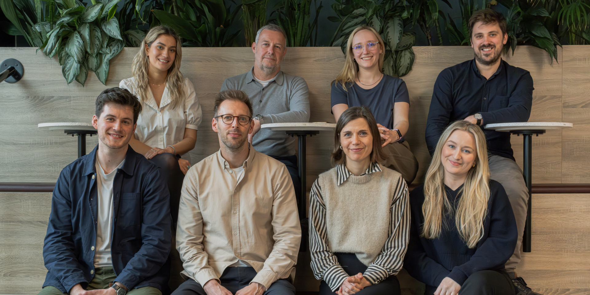 A group of nine diverse young professionals posing together in an indoor setting with green plants and wooden decor.