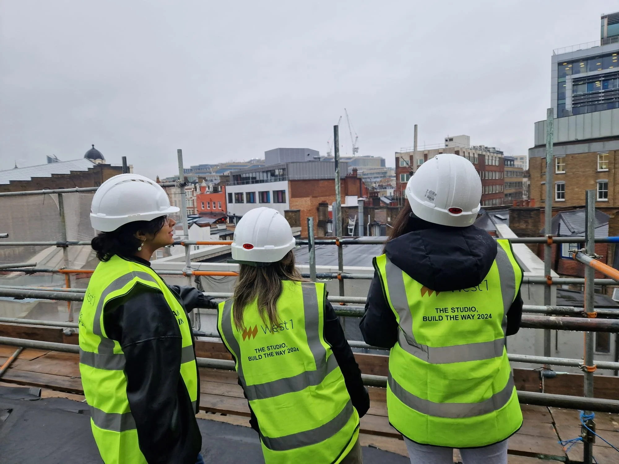 Three construction workers wearing yellow safety vests and white hard hats standing on a rooftop, overlooking a city skyline with various buildings and cranes under an overcast sky.