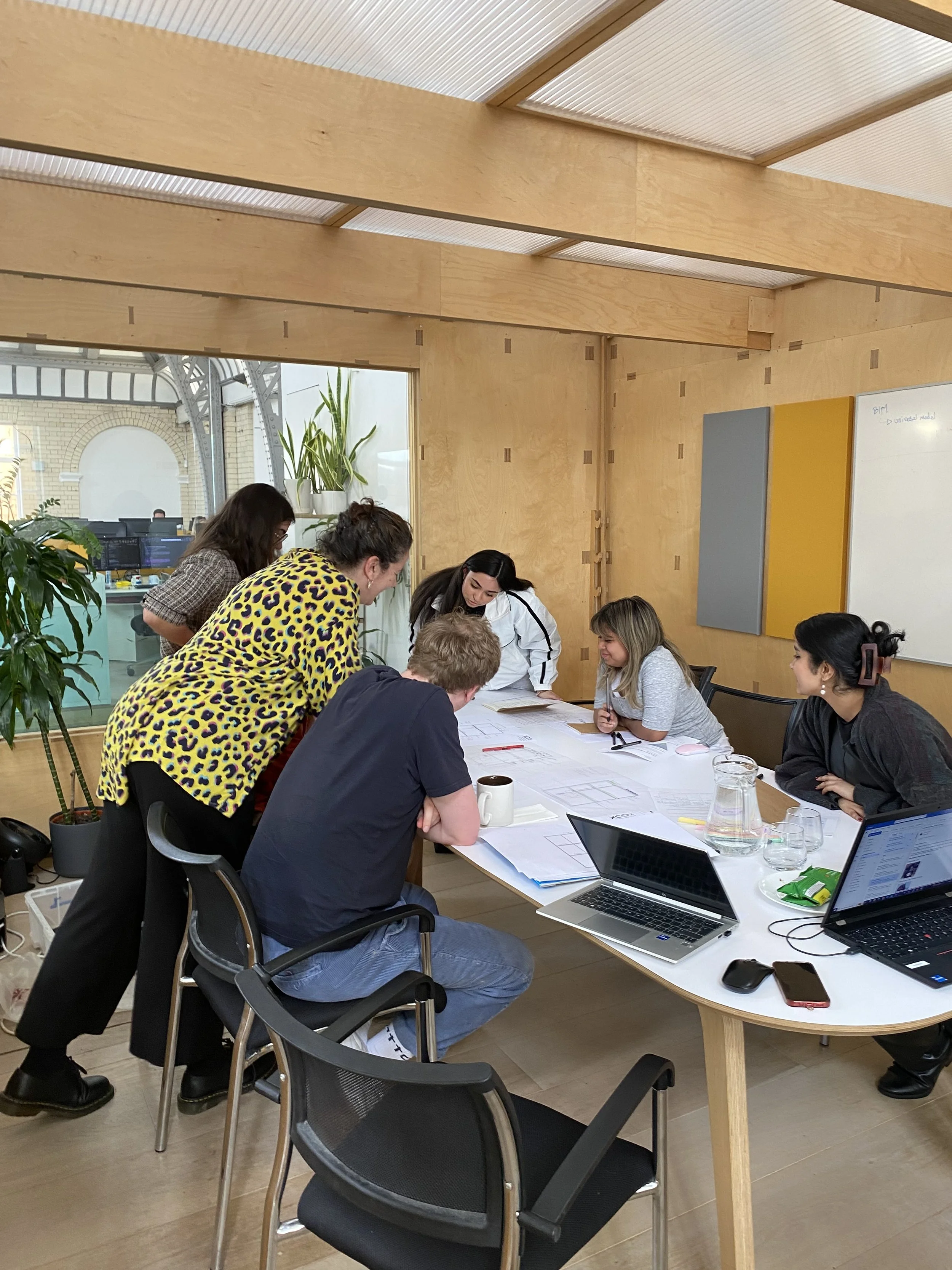 People gathered around a long conference table during a meeting or workshop in a well-lit room with wooden walls and large windows, with laptops, notebooks, and drinks on the table.
