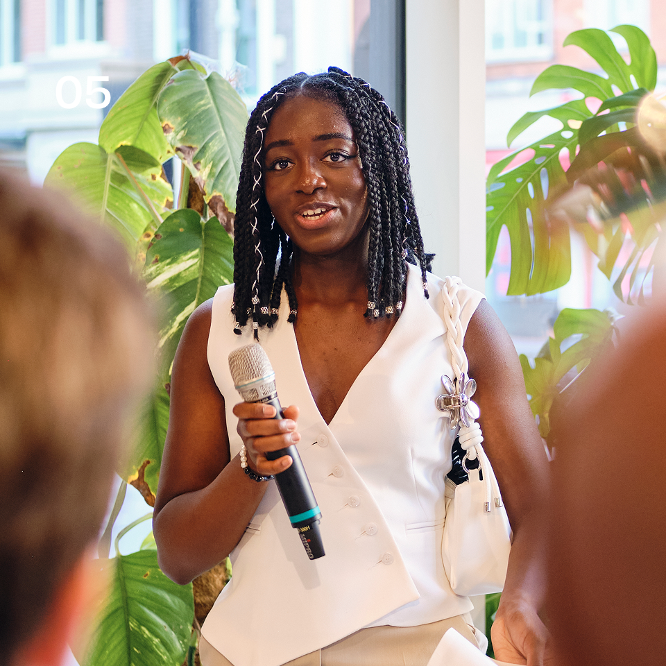 Woman with braided hair speaking into a microphone in a room with large green plants and windows.