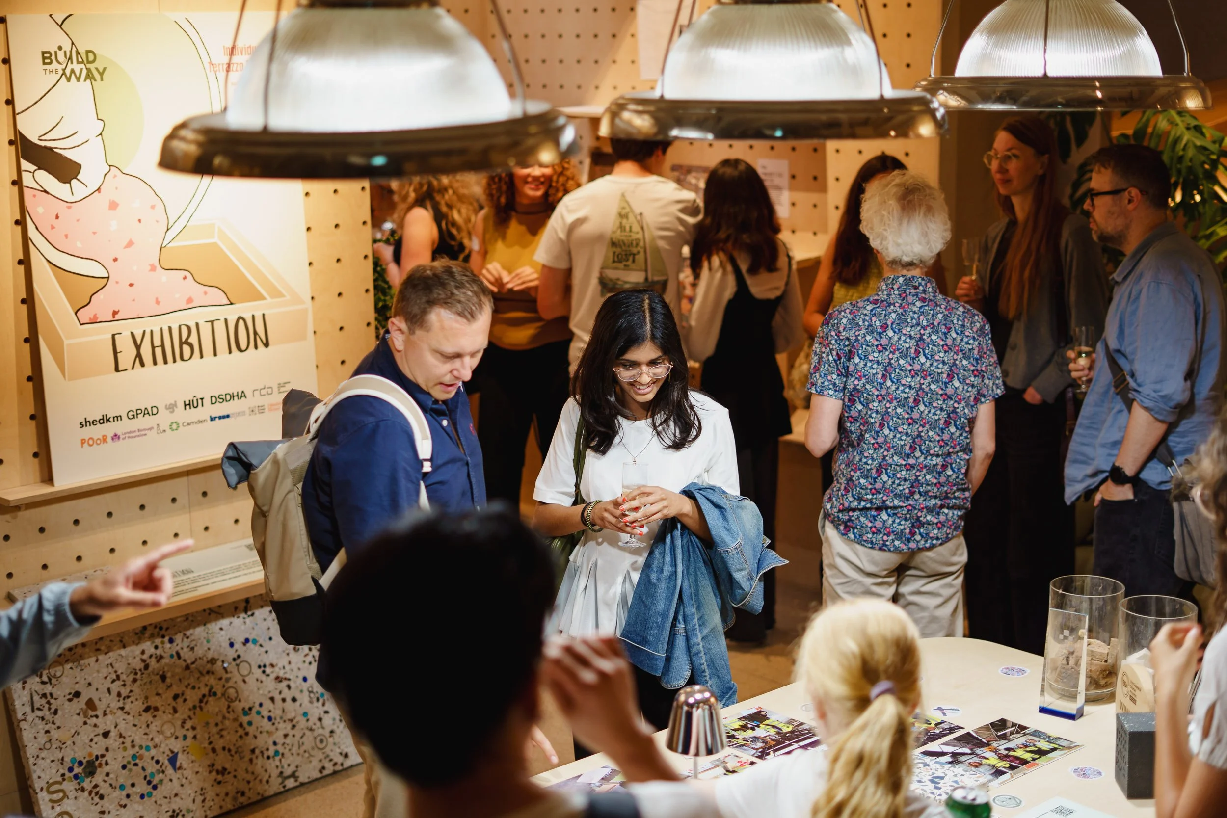 A gallery opening with people socializing, some holding drinks. A large sign with the word 'Exhibition' and artwork can be seen on the wall. The space has warm lighting and a wooden wall background.