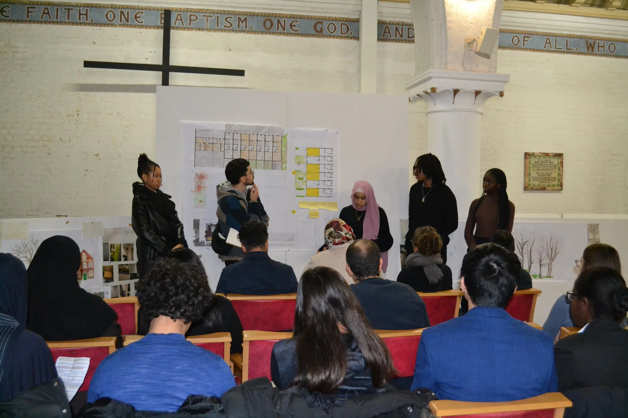 A group of five people stands in front of a white presentation board with diagrams inside a church, with the congregation seated facing them. The church has a large cross, brick walls painted white, and a banner with the words 'Faith, One Baptism, One God' across the top.