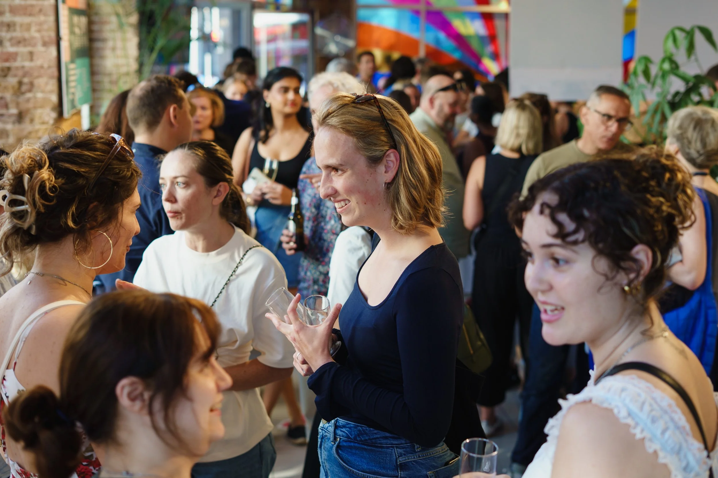 People gathered at an indoor event, socializing and holding drinks, with colorful artwork and a brick wall in the background.
