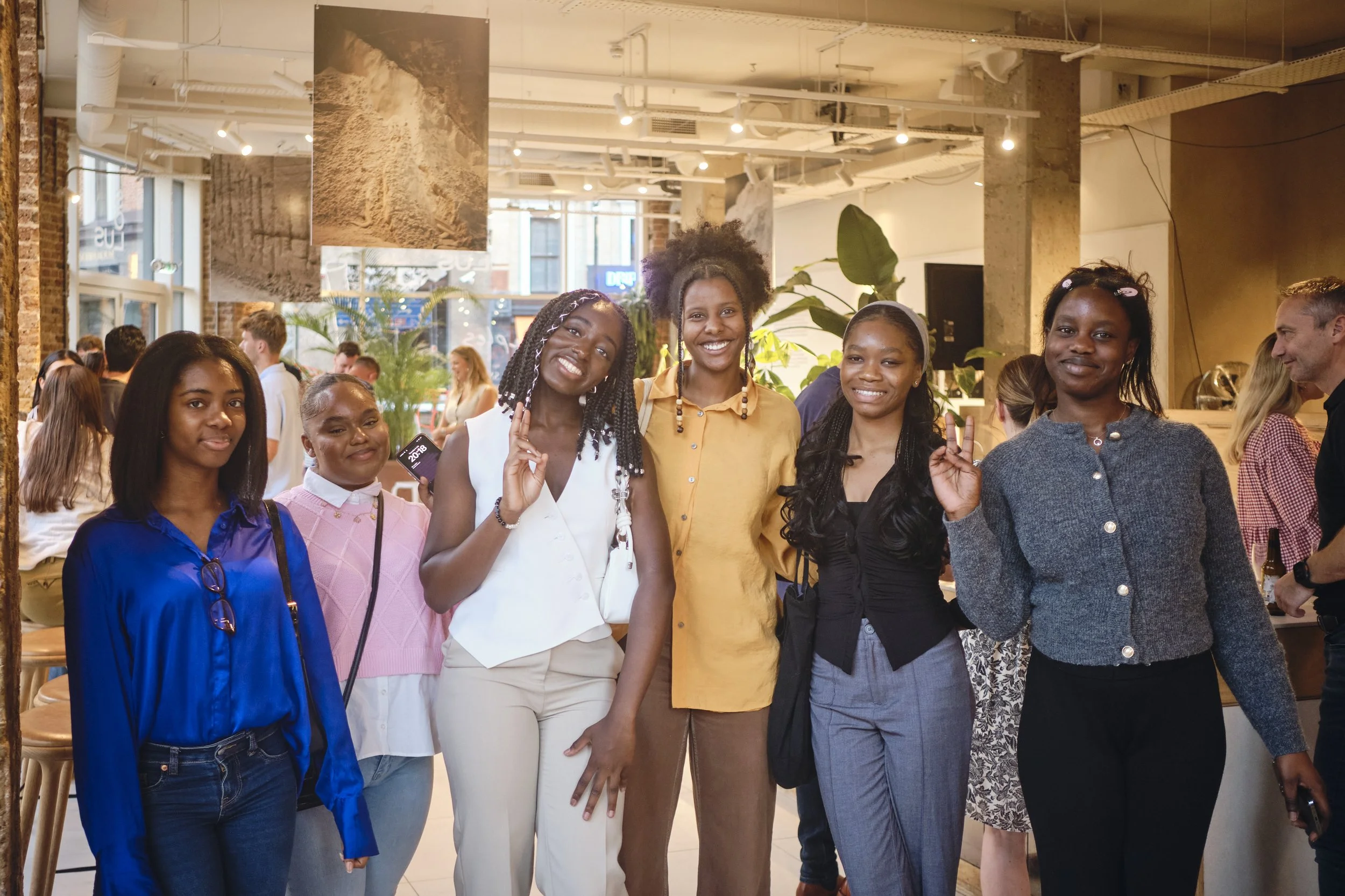 Group of six diverse women smiling and posing together inside a modern art gallery or cafe with brick walls, large windows, hanging photographs, and plants.