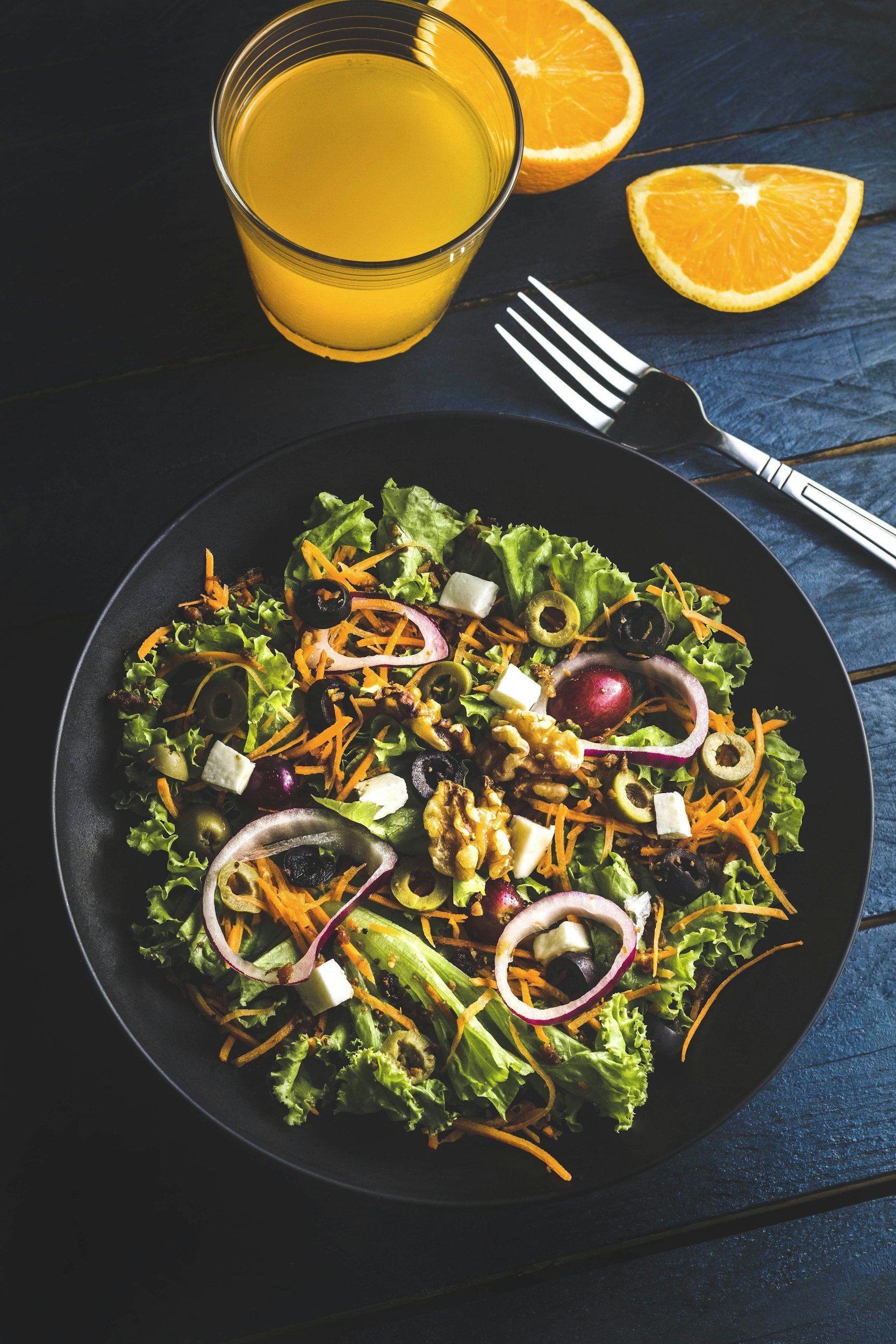 Bunter Salat mit grünen Blättern, Tomaten, roten Zwiebeln, Oliven, Käse und Nüssen auf einem schwarzen Teller. Daneben liegt ein Glas Orangensaft und eine halbierte Orange. Ein Messer und eine Gabel sind auf einem dunklen Holztisch.