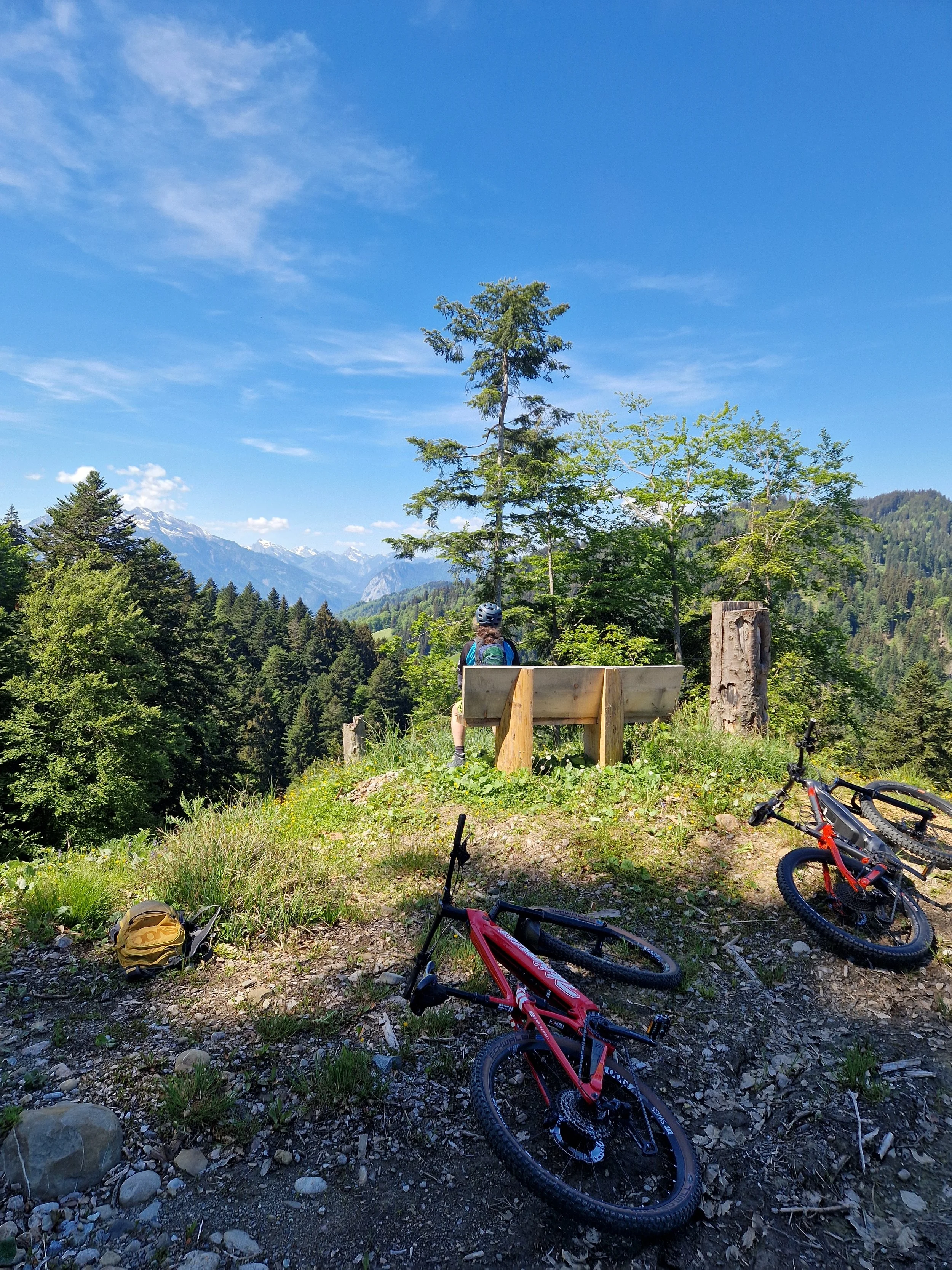 Mountainbikes liegen auf dem Boden, ein Rucksack ist sichtbar, im Hintergrund sind Bäume und Berge, der Himmel ist blau und es ist sonnig.