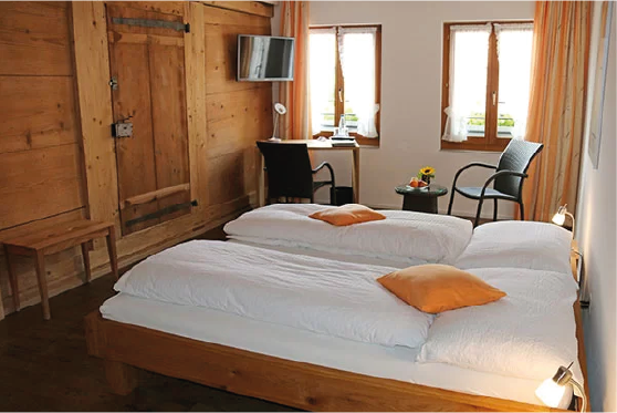 Ein Hotelzimmer mit zwei Einzelbetten, einem kleinen Tisch, zwei Stühlen und einem Fernseher, natürliche Lichtquellen durch zwei Fenster, Holzverkleidung an den Wänden.