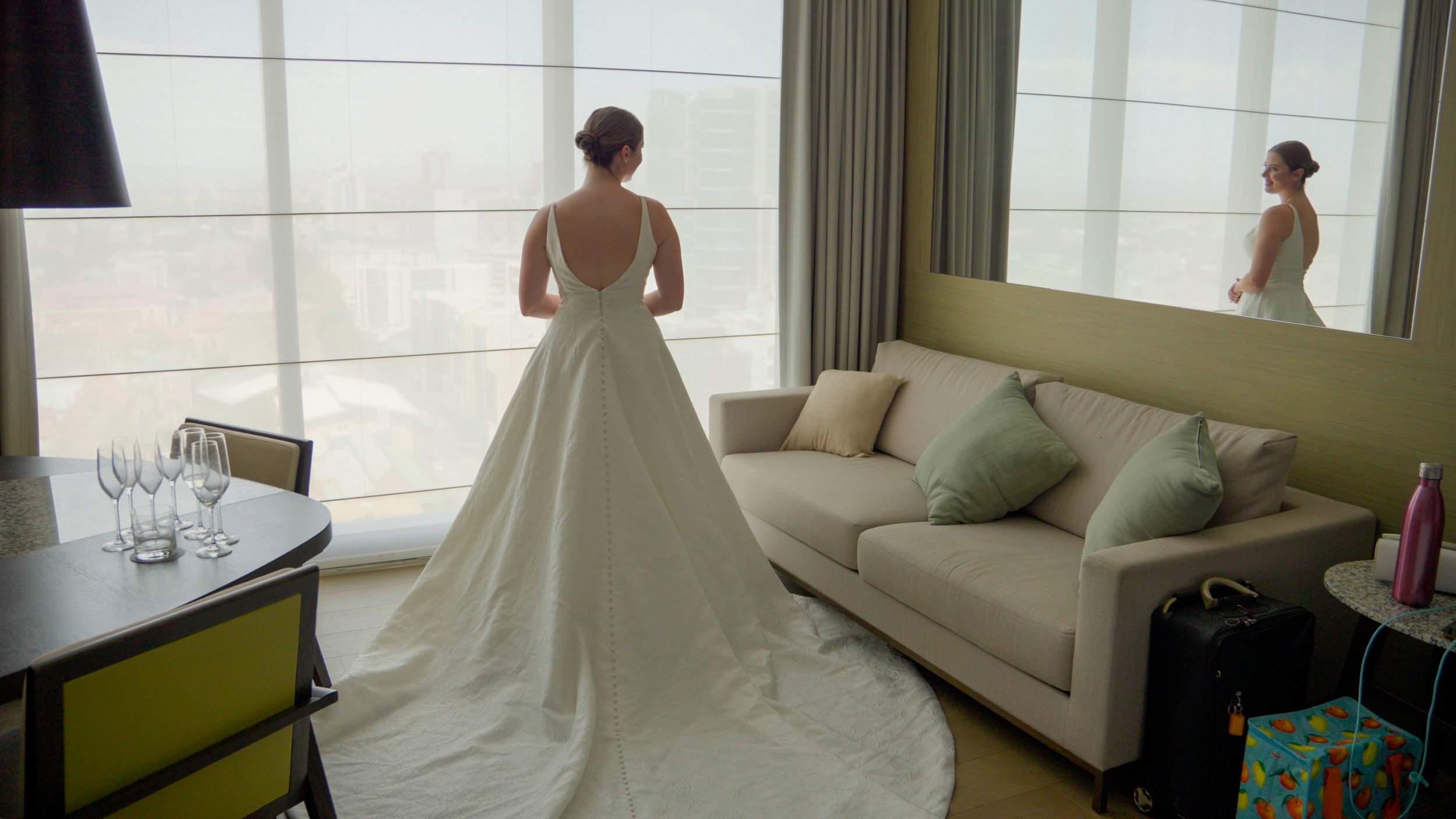 A woman in a wedding dress stands near a large window. She has her back to the camera, and her reflection is visible in a wall mirror. The room is elegantly furnished with a sofa, green cushions, a small table, and a few glasses placed on a table.
