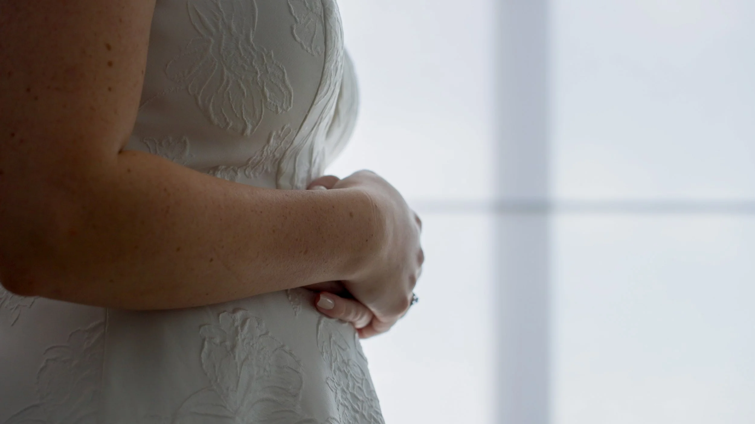 Close-up of a person in a white floral-patterned dress, hands clasped at the front.