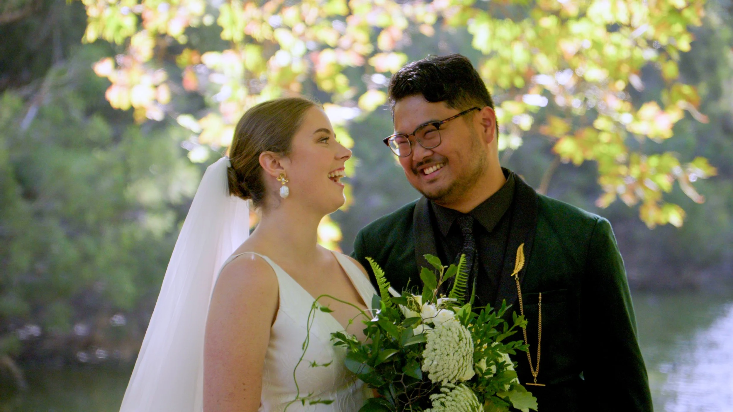 Bride and groom smiling at each other, holding a bouquet, with greenery in the background.