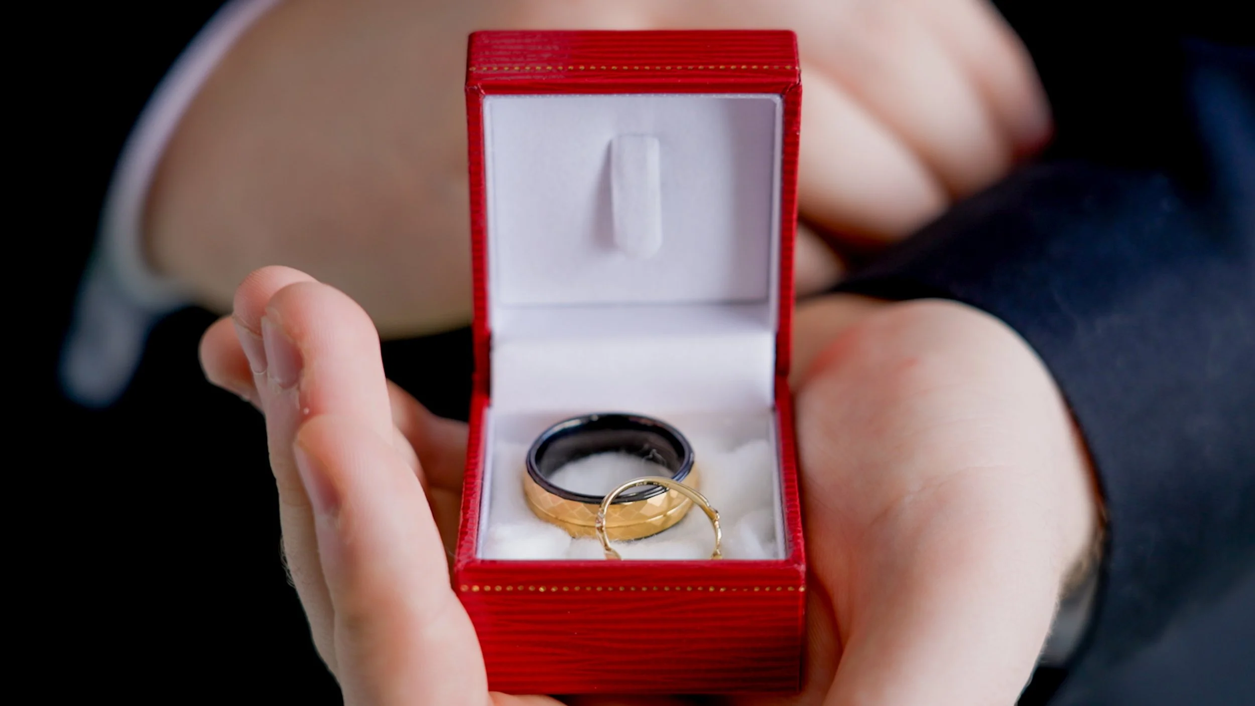 Two rings in a red jewelry box held by a person wearing a suit; one ring is black and gold, the other is a gold band.