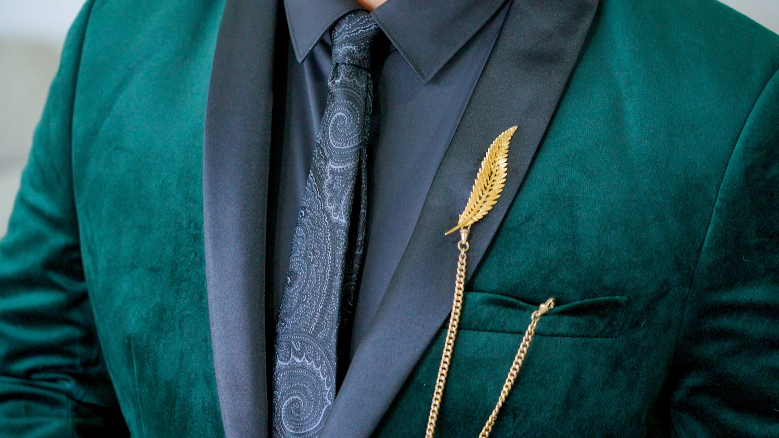Man wearing a velvet green suit with a black, paisley-patterned tie and a golden leaf lapel pin with a chain.