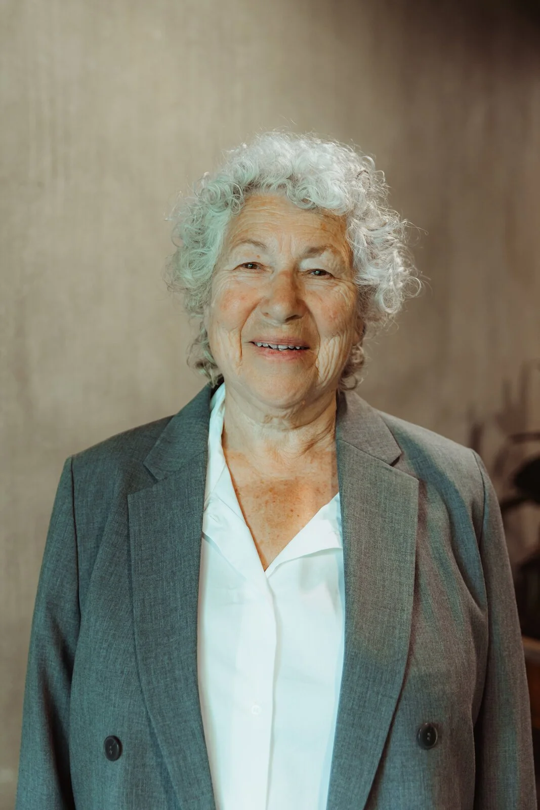 Christy, Betwixt dancer, smiling gently in a grey blazer with short white curly hair.