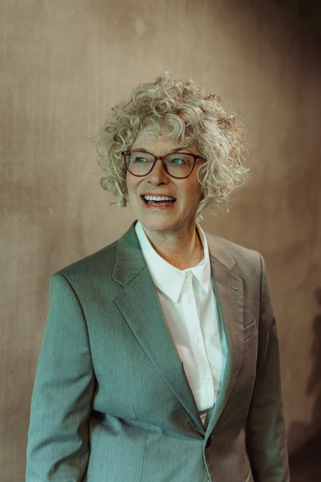 Penny, Betwixt dancer, laughing in a teal-grey blazer with curly grey hair and glasses.