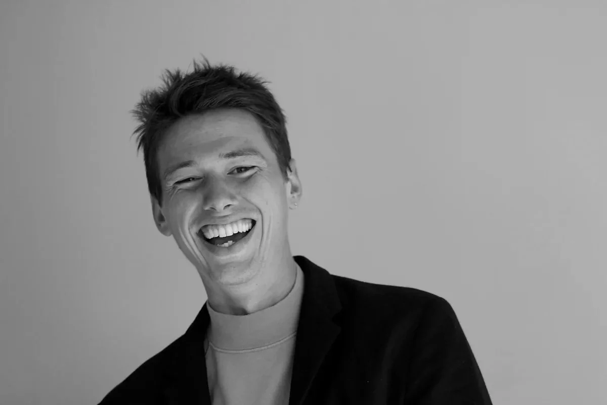 Young man with short hair, smiling, in light shirt and dark jacket against plain background.