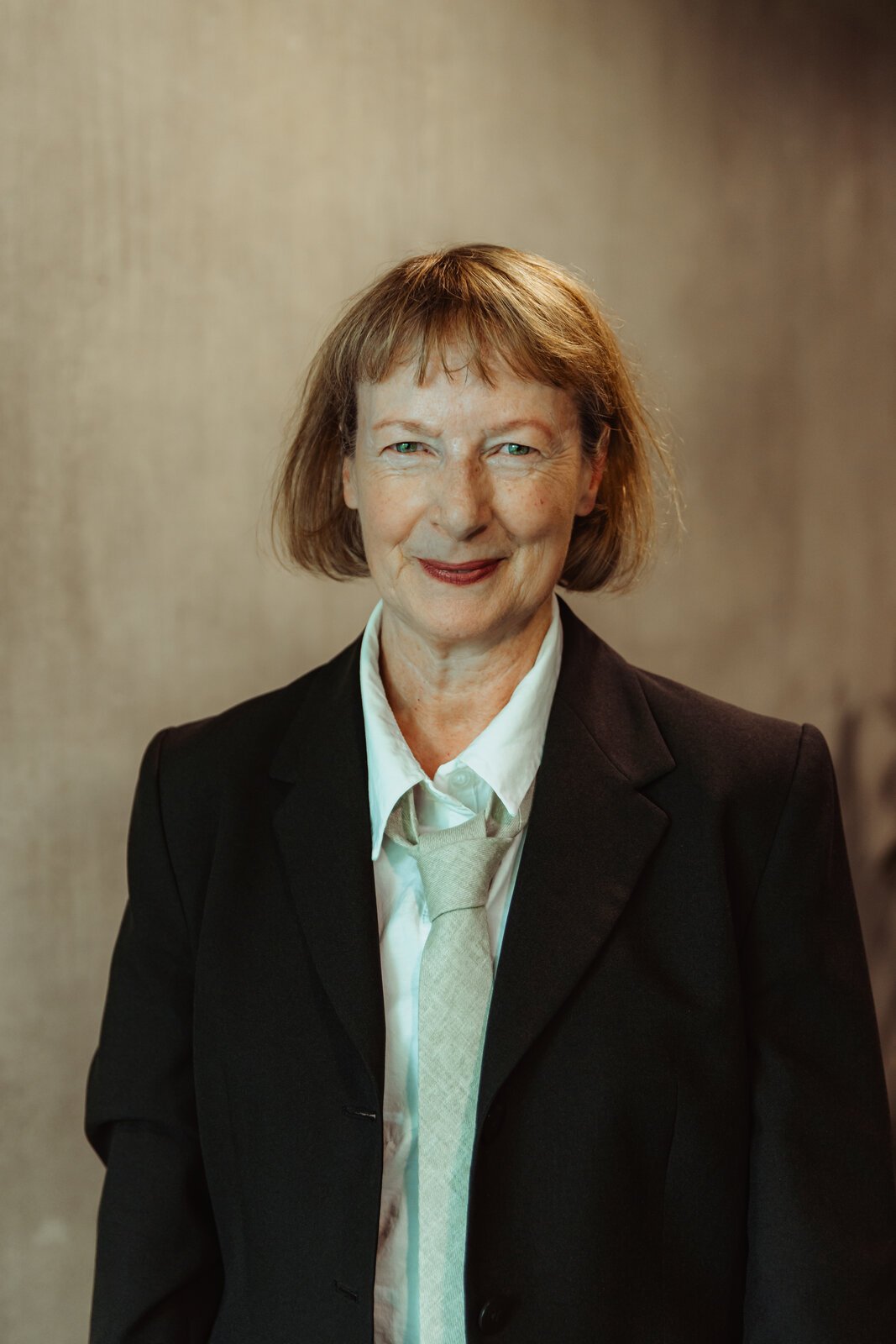 Betwixt cast member with an auburn bob and fringe, smiling in a black blazer with a sage green tie.