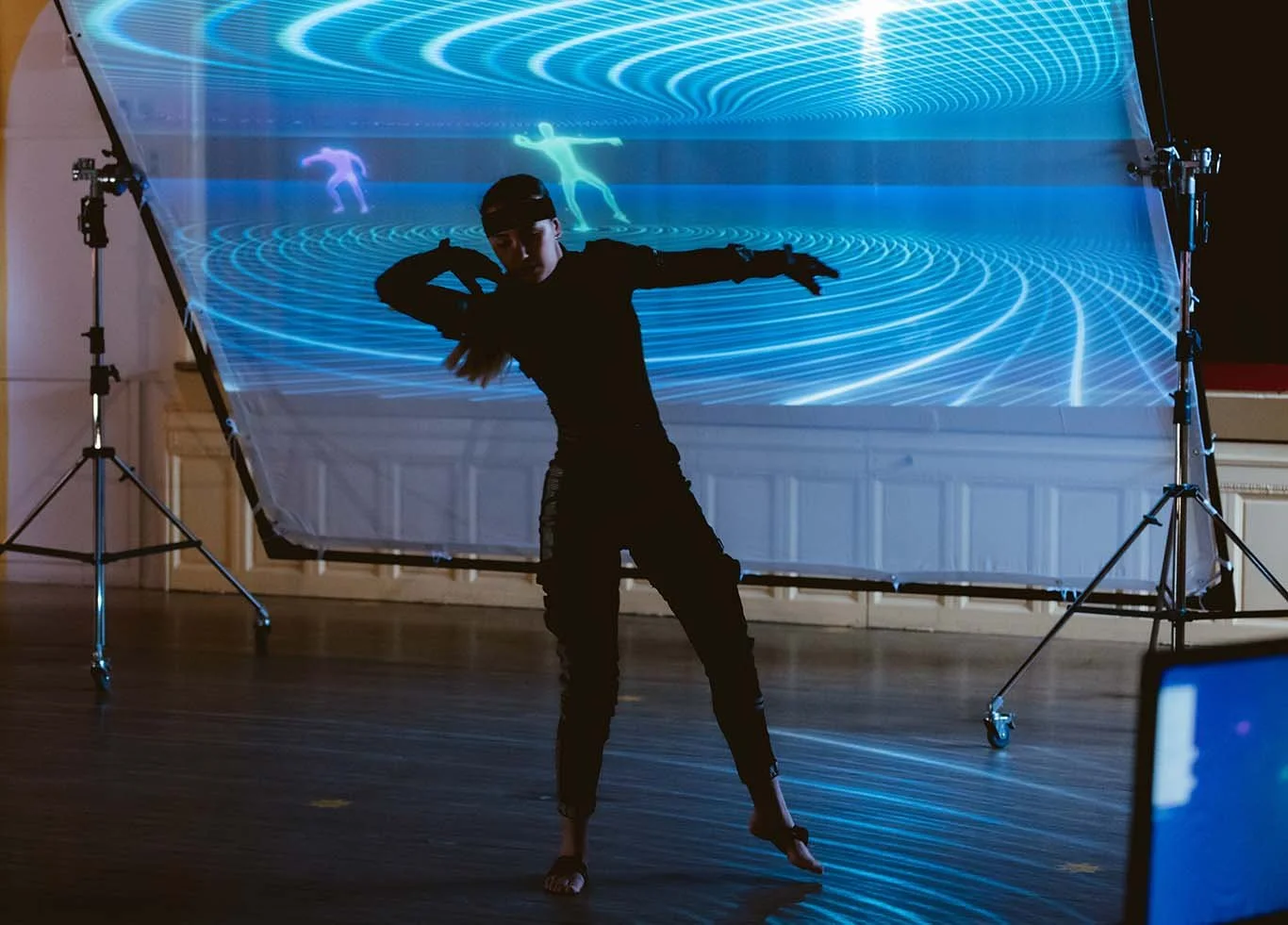 A dancer performs in front of a digital projection with swirling blue patterns and holographic figures, creating a futuristic stage setting.