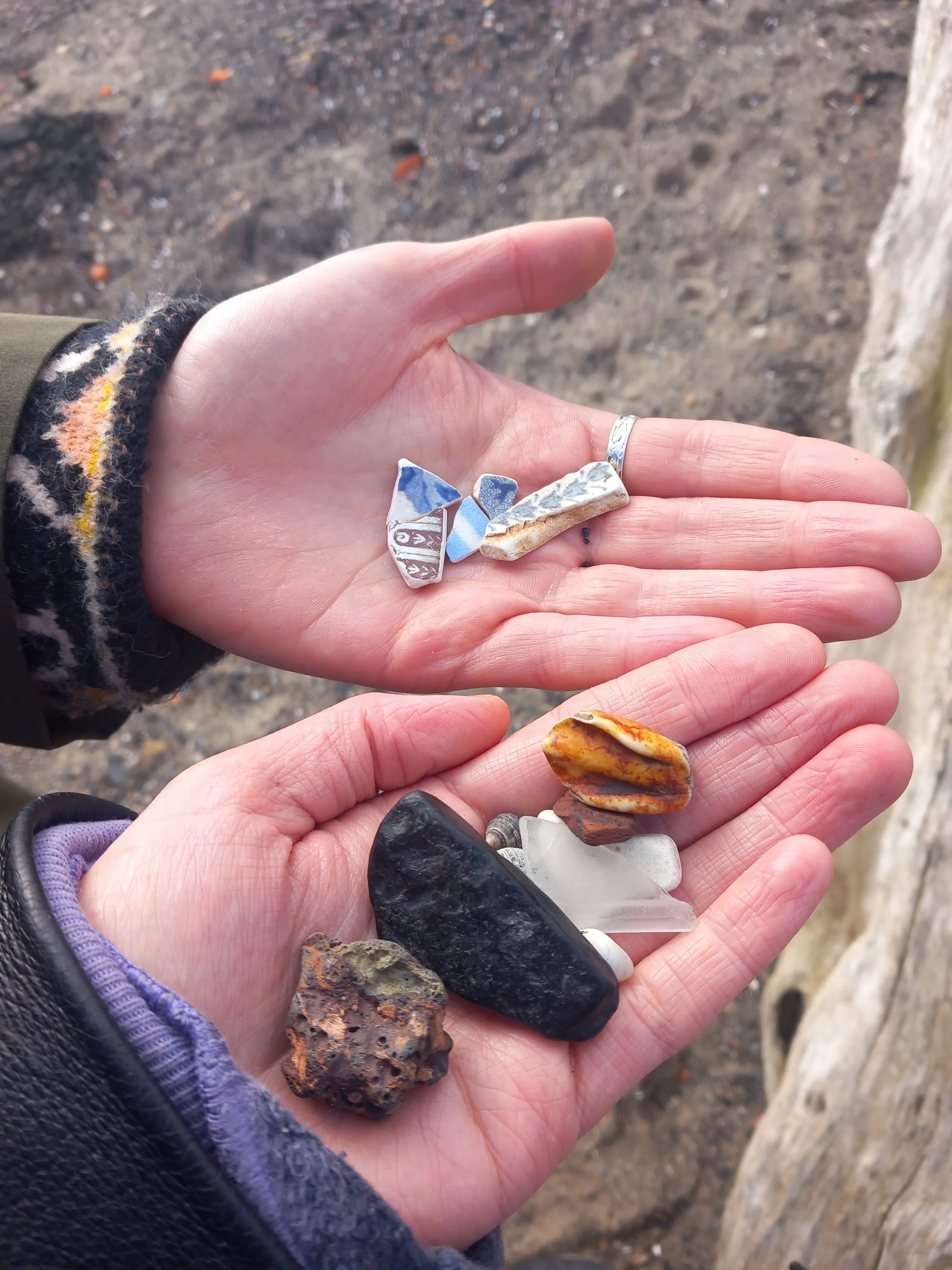 close up of two people's hands with pale skin holding out things they have found on the beach, including pieces of pottery, rocks, and sea glass, with sand in the background.