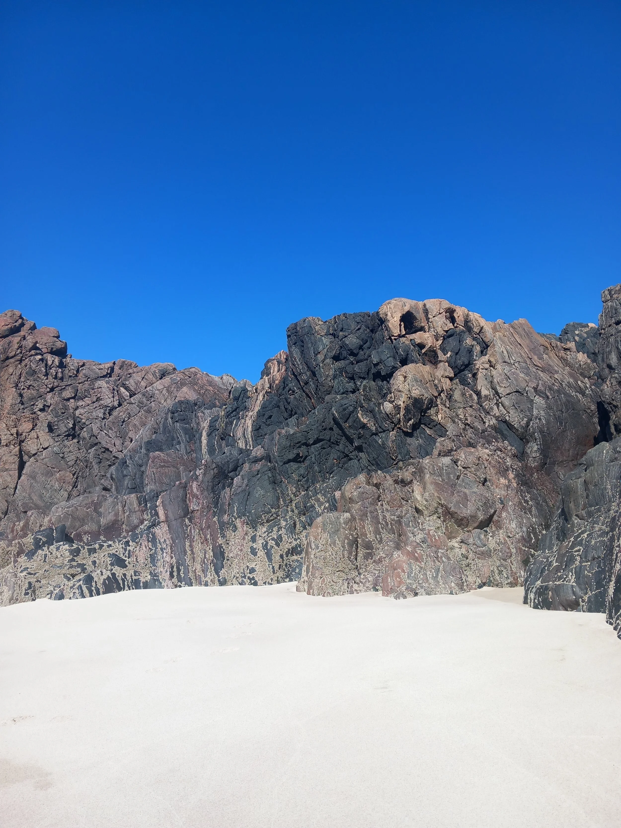 brown and black rocks with some barnacles attached sit on a white sandy beach with a clear bright blue sky above