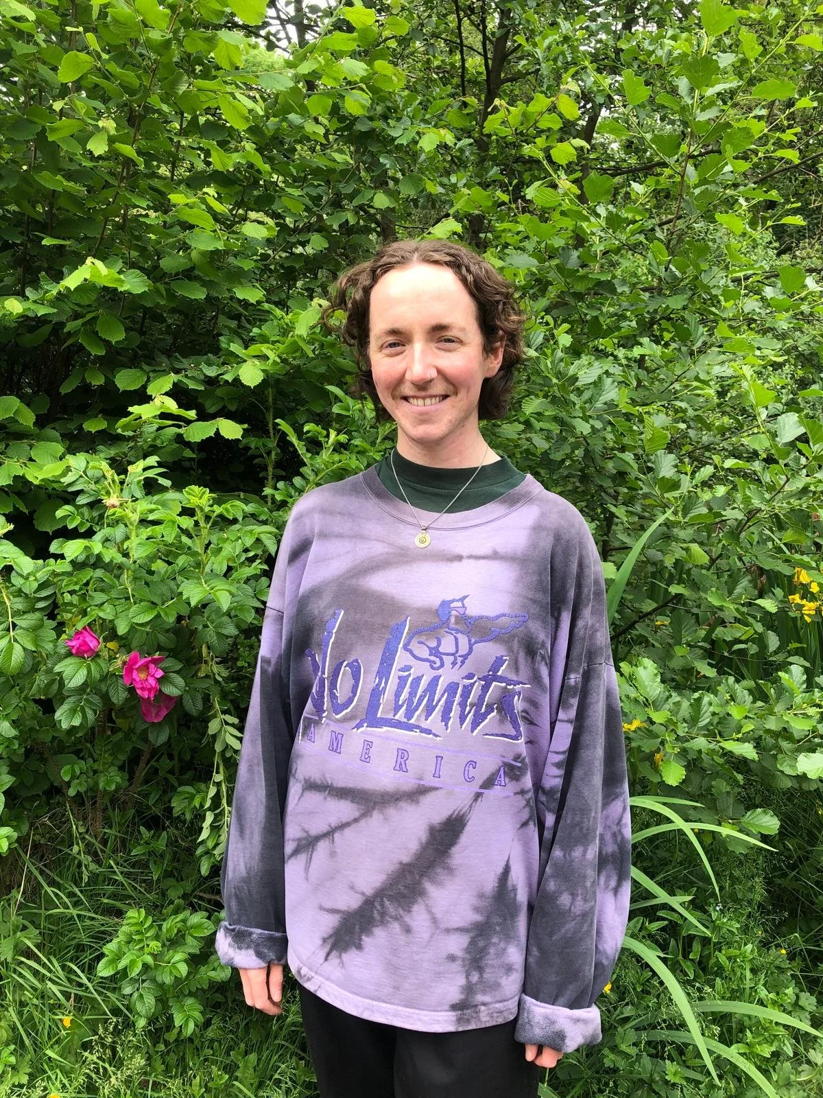 a picture of River, a white person with short brown curly hair and a purple jumper, stands smiling in front of some green bushes with two pink roses to their left
