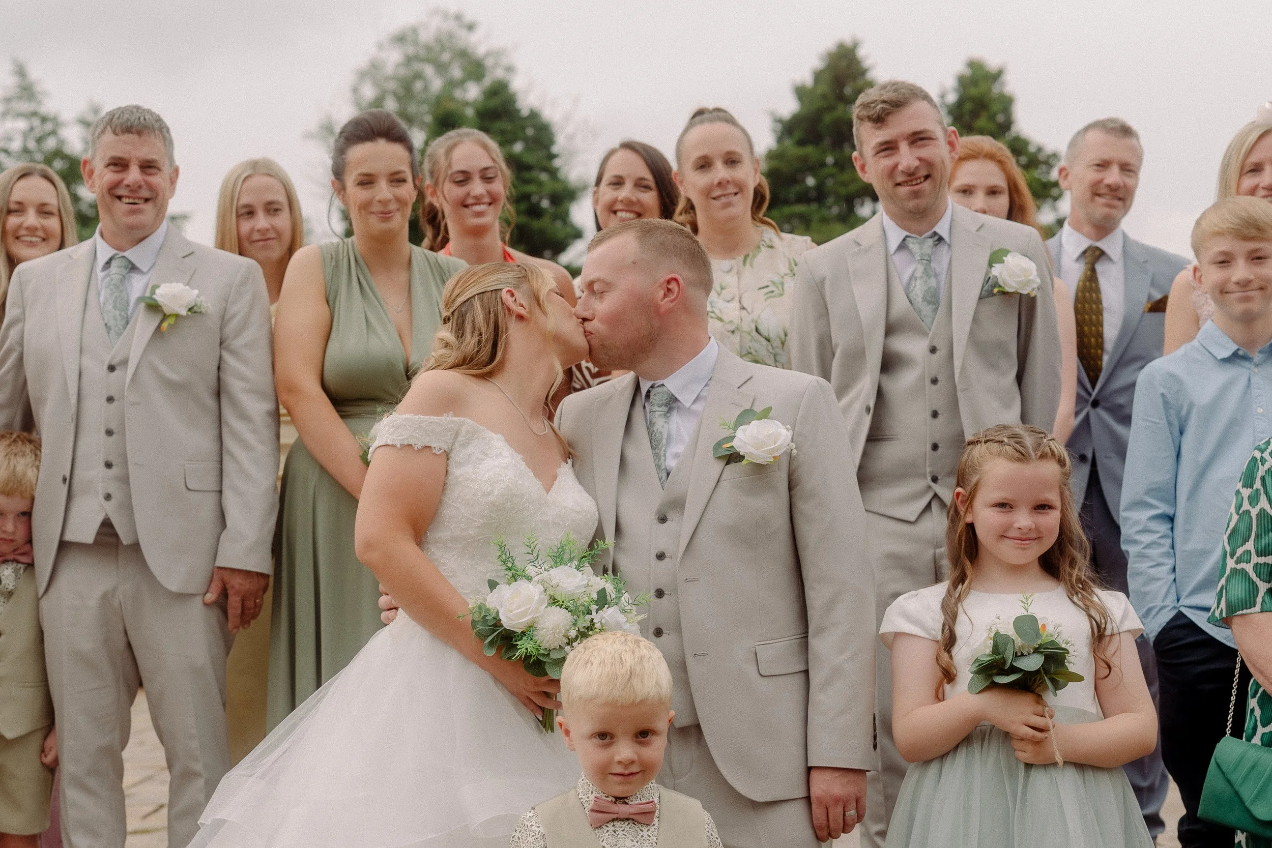 Bride and bridesmaids smiling with bouquets, wearing peach and blush dresses outdoors, with greenery in the background.